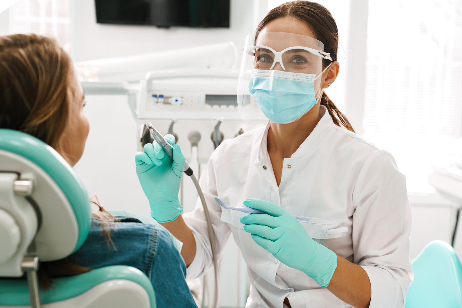 A hygienist in a face shield holding a polisher and a toothbrush beside a patient in the chair