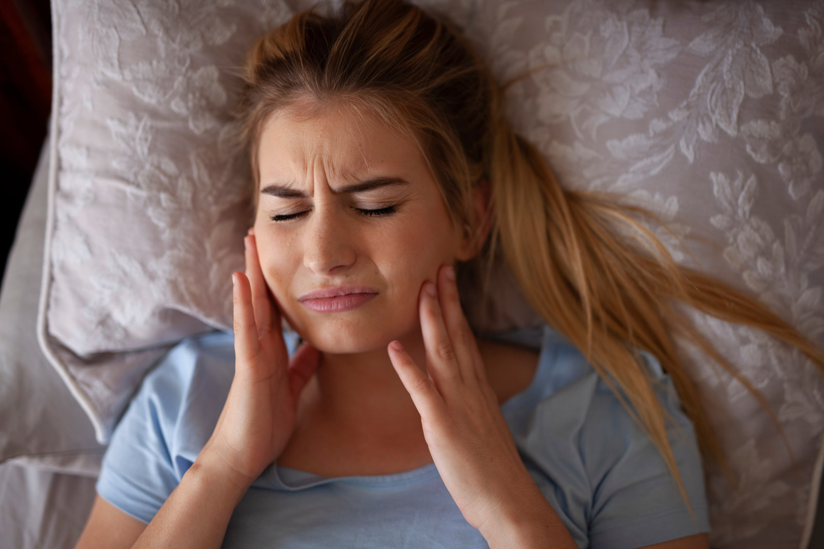 A woman lying in bed pressing both sides of her jaw in discomfort