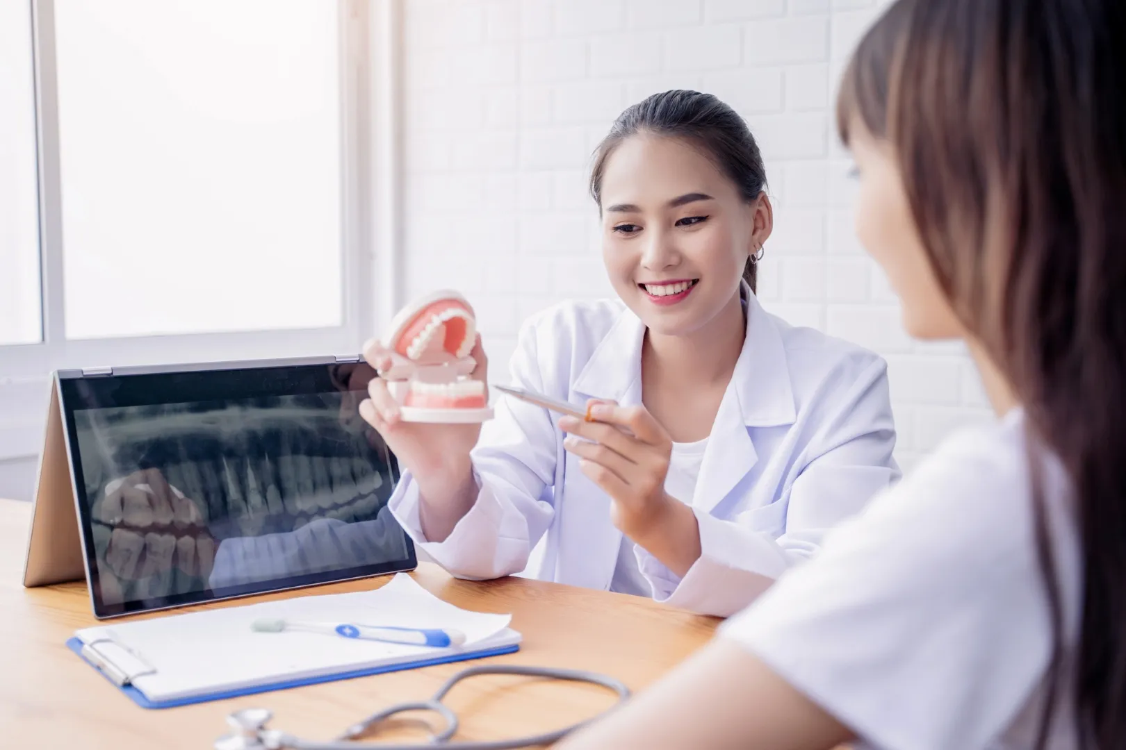 A dentist showing a patient a model of the teeth alongside an X-ray on a laptop