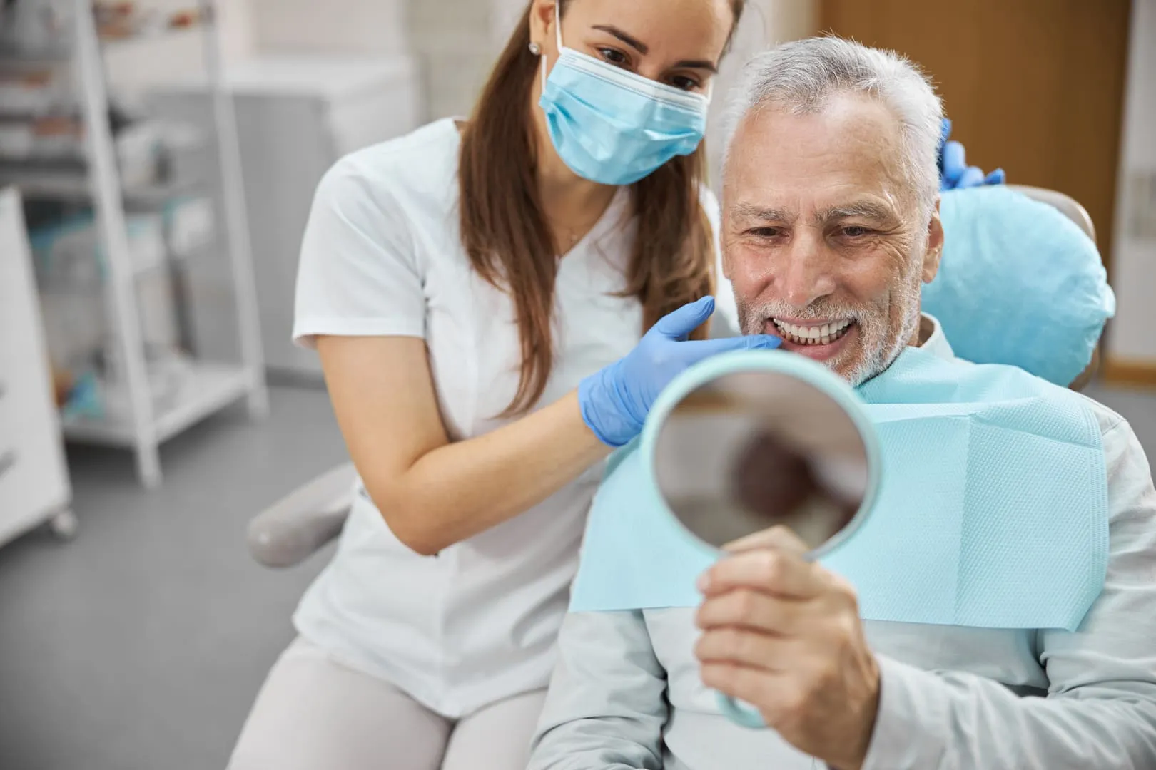 An older man looking at his teeth in a hand mirror while a dental assistant points them out