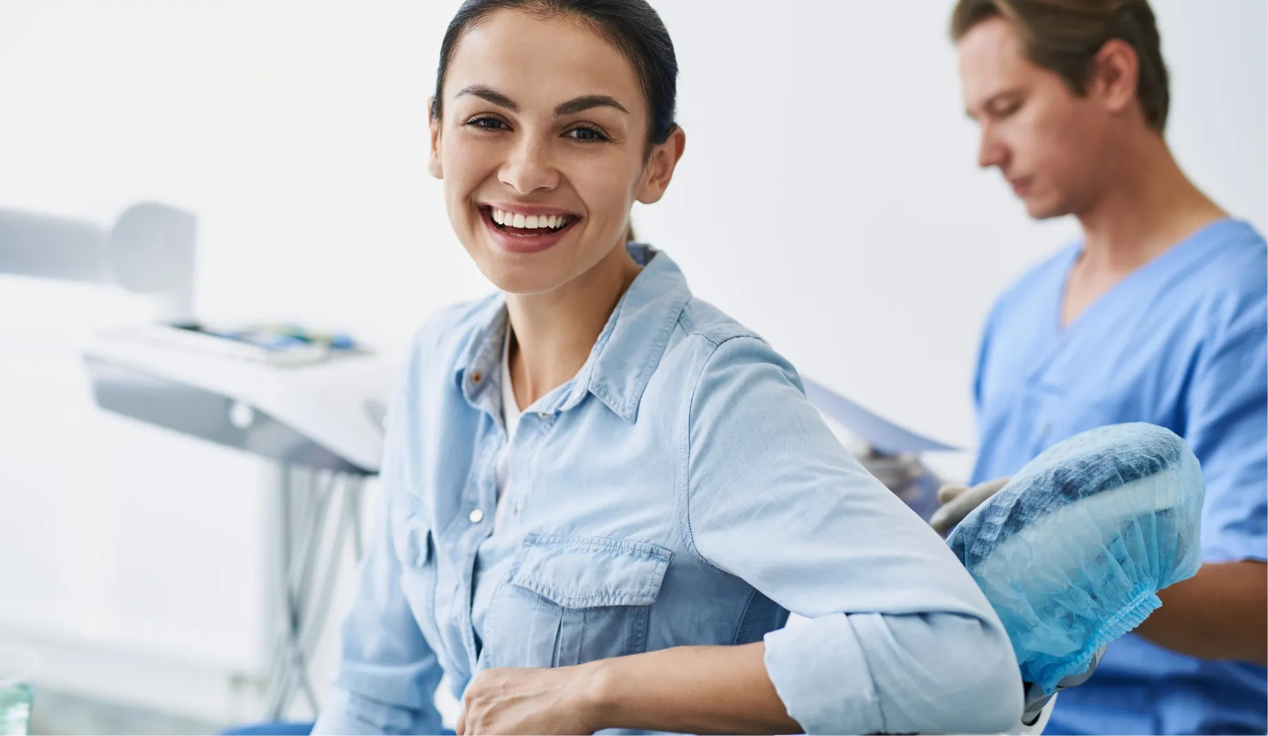 A smiling patient seated in the treatment room, with a clinician working behind her